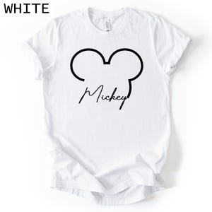May include: White t-shirt with a black outline of Mickey Mouse ears and the name "Mickey" in a cursive font. The word "WHITE" is printed in the upper left corner. The shirt is made of a soft material.