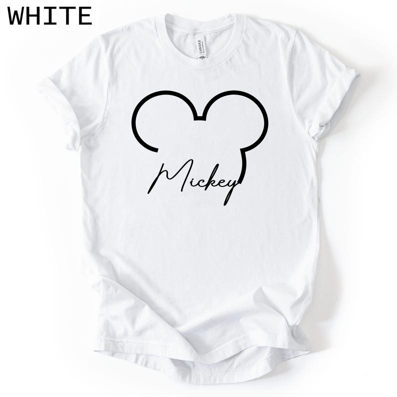 May include: White t-shirt with a black outline of Mickey Mouse ears and the name "Mickey" in a cursive font. The word "WHITE" is printed in the upper left corner. The shirt is made of a soft material.