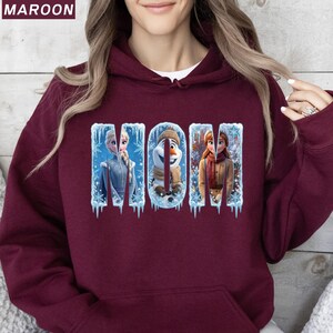 Puede incluir: Sudadera con capucha de color granate con la palabra "MOM" en un dise&ntilde;o de invierno. Las letras presentan im&aacute;genes de los personajes de la pel&iacute;cula Frozen: Elsa, Olaf y Anna.