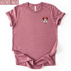 May include: A heather mauve short-sleeve t-shirt featuring a small graphic of Minnie Mouse on the left chest. The character is depicted with her signature red and white polka dot bow, white gloves, and black ears, centered on the upper chest area.
