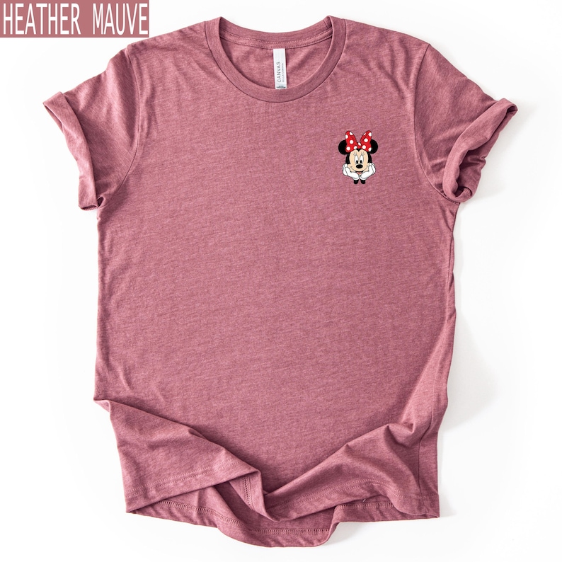 May include: A heather mauve short-sleeve t-shirt featuring a small graphic of Minnie Mouse on the left chest. The character is depicted with her signature red and white polka dot bow, white gloves, and black ears, centered on the upper chest area.