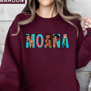 May include: A maroon sweatshirt with the word "MOANA" in turquoise, featuring cartoon character illustrations. The sweatshirt is a solid maroon colour, with the text and graphics in the centre.