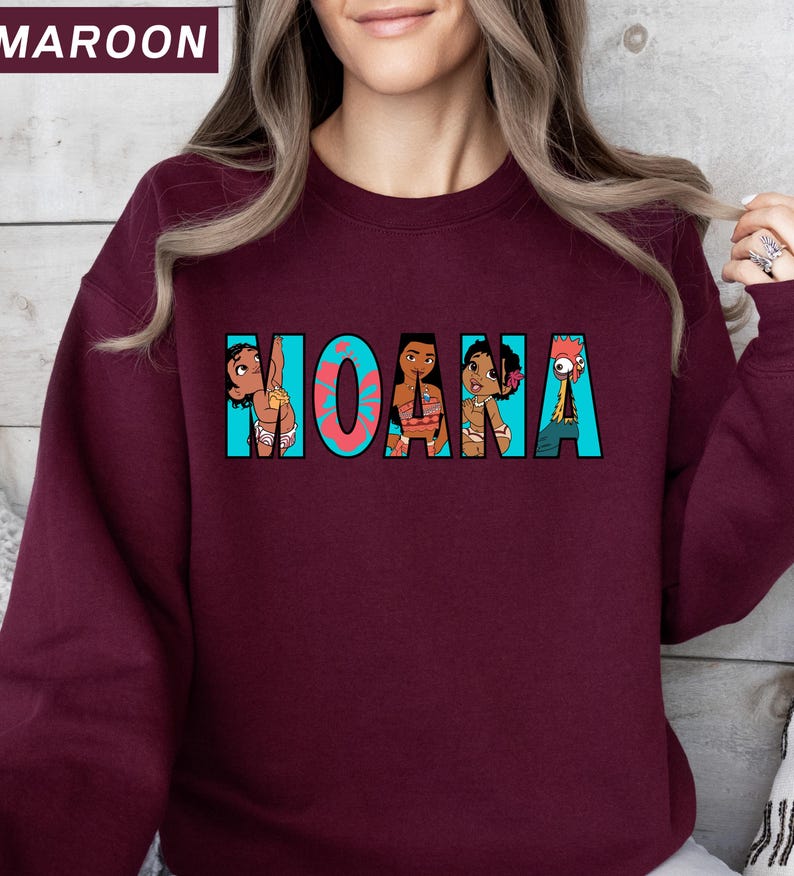 May include: A maroon sweatshirt with the word "MOANA" in turquoise, featuring cartoon character illustrations. The sweatshirt is a solid maroon colour, with the text and graphics in the centre.