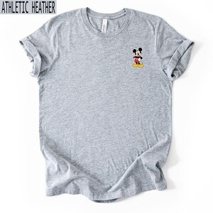 May include: An athletic heather grey short-sleeved t-shirt featuring a small graphic of Mickey Mouse on the left chest. The character is depicted in his classic red shorts and yellow shoes. The shirt has a crew neckline and a standard fit.