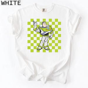 May include: White t-shirt featuring a cartoon illustration of Buzz Lightyear from Toy Story. The character is depicted in his signature space ranger suit against a green and white checkered background. The word "WHITE" is printed at the top.