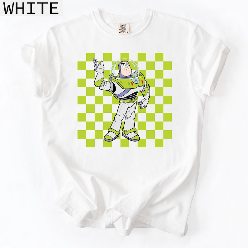 May include: White t-shirt featuring a cartoon illustration of Buzz Lightyear from Toy Story. The character is depicted in his signature space ranger suit against a green and white checkered background. The word "WHITE" is printed at the top.