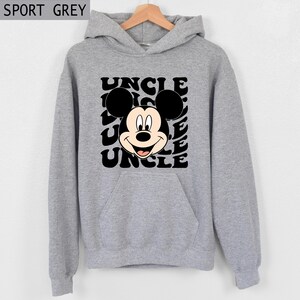 Pu&ograve; includere: Felpa con cappuccio grigio sportivo con una grafica di Topolino e la parola "UNCLE" in un motivo ripetuto. La felpa ha una tasca frontale e un cappuccio con coulisse. Il testo "SPORT GREY" &egrave; nell'angolo in alto a sinistra.