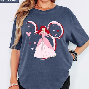 May include: Denim blue t-shirt featuring a graphic of a princess in a pink dress, with red hair, inside a Mickey Mouse ear design. Pink balloons, stars, and hearts surround the princess. The word "DENIM" is in the upper left corner.