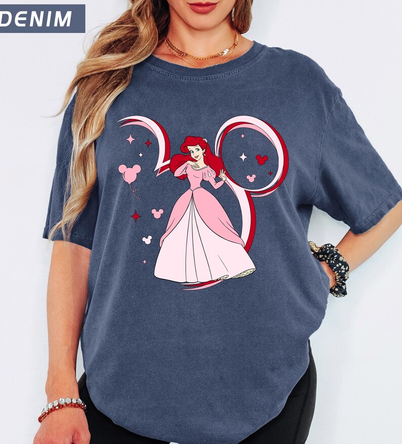 May include: Denim blue t-shirt featuring a graphic of a princess in a pink dress, with red hair, inside a Mickey Mouse ear design. Pink balloons, stars, and hearts surround the princess. The word "DENIM" is in the upper left corner.