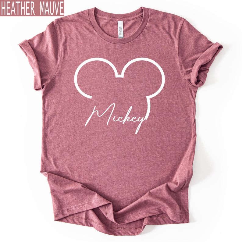 May include: Heather mauve t-shirt featuring a white Mickey Mouse outline and the name "Mickey" in cursive. The shirt has a crew neck and is made of a soft material. The top left corner of the image has the text "HEATHER MAUVE".