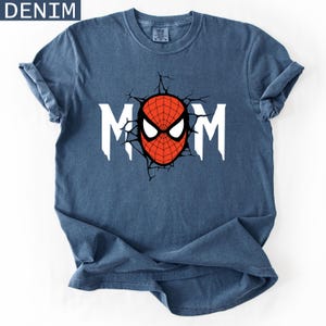 May include: A denim blue short-sleeve t-shirt featuring a graphic print on the chest. The design displays the white text MOM with a red and black spider-themed mask replacing the letter O, surrounded by a black cracked wall effect graphic.