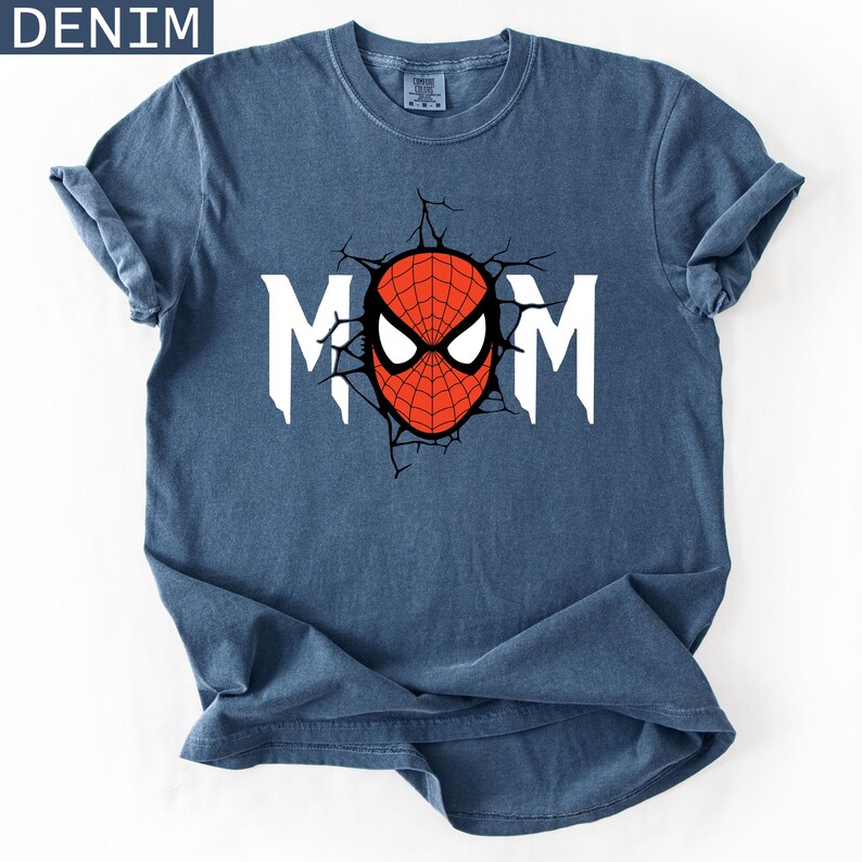 May include: A denim blue short-sleeve t-shirt featuring a graphic print on the chest. The design displays the white text MOM with a red and black spider-themed mask replacing the letter O, surrounded by a black cracked wall effect graphic.