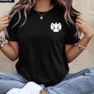 May include: Black t-shirt with a white graphic of a stylized eagle emblem on the chest. The word "BLACK" is in a black rectangle in the upper left corner. The shirt is worn with blue jeans and white sneakers.