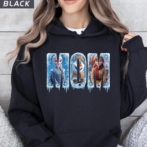 Puede incluir: Sudadera con capucha negra con la palabra "MOM" en letras azul hielo. Las letras presentan im&aacute;genes de personajes de la pel&iacute;cula Frozen. La sudadera es de color oscuro.