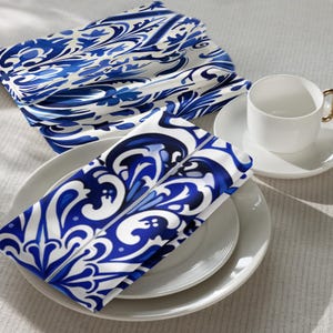 Op de afbeelding: Witte kop en schotel met een gouden handvat, naast gevouwen servetten met een blauw-wit patroon op witte borden. De servetten hebben een gedetailleerd blauw bloemmotief.