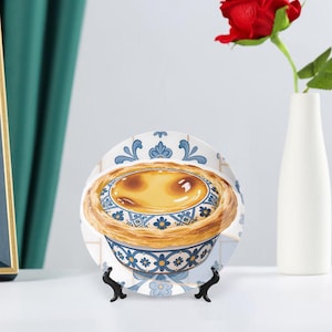 May include: A decorative plate featuring a custard tart design. The plate has a white background with blue floral patterns and a detailed image of the dessert. It is displayed on a black stand, with a red rose in a white vase in the background.