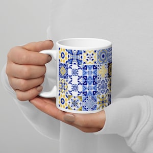 May include: White ceramic mug featuring a patchwork design of blue and yellow geometric patterns. The mug has a standard handle and a glossy finish, perfect for hot beverages. The design is reminiscent of traditional tilework.