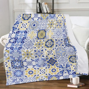 Può includere: Una coperta decorativa con un design patchwork. La coperta presenta un motivo ripetuto di piastrelle blu e gialle su sfondo bianco. Le piastrelle hanno vari disegni floreali e geometrici.