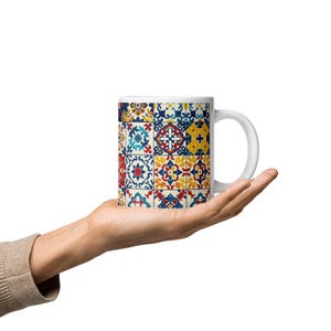 May include: White ceramic mug featuring a colorful tile pattern in shades of blue, yellow, red, and orange. The mug has a standard handle and a smooth, glossy finish. The design is reminiscent of traditional tilework.