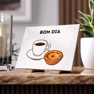 Könnte beinhalten: Eine weiße Keramikfliese mit den Worten "BOM DIA" in Schwarz. Die Fliese zeigt eine Zeichnung einer Kaffeetasse, Untertasse und eines Gebäckstücks. Die Fliese wird von zwei kleinen Holzblöcken getragen. Eine Kerze und eine Pflanze sind im Hintergrund.
