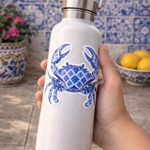 May include: A white water bottle with a silver lid and handle, featuring a blue and white crab sticker. The crab design has a detailed, patterned appearance. The background includes a blue and white tile pattern.