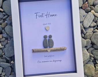 May include: A framed artwork with a white border, featuring the text "First Home" and "April 2025". Two pebble figures sit on a wooden branch, with a heart above and below. The names "Jack and Becca" and the phrase "New memories new beginnings" are also included.