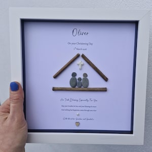 May include: Framed artwork with a white border. The artwork features a house design made of sticks and pebbles, with a cross and family figures. Text includes "Oliver" and "On your Christening Day 7th March 2016".