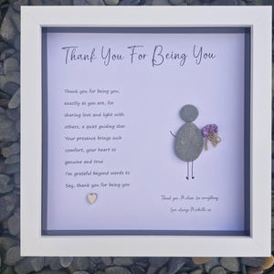 May include: A framed artwork with the text "Thank You For Being You." The artwork features a pebble art figure holding a bouquet of purple flowers. A small wooden heart is at the bottom. The frame is white.