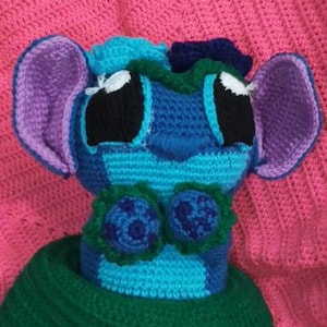 May include: A crocheted plush toy featuring a blue and light blue head with large black eyes, purple-lined ears, and a dark blue nose. The figure has two dark blue circular accents on the chest and is wearing a solid dark green crocheted base.