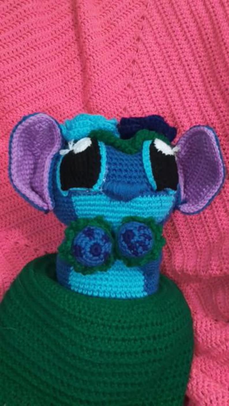 May include: A crocheted plush toy featuring a blue and light blue head with large black eyes, purple-lined ears, and a dark blue nose. The figure has two dark blue circular accents on the chest and is wearing a solid dark green crocheted base.