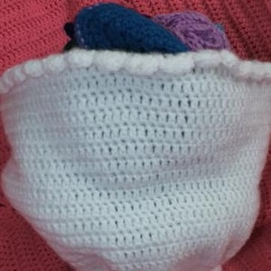 May include: A bouquet of crocheted flowers wrapped in a white crocheted paper-style sleeve with a scalloped edge. The flowers feature various colors including dark blue and light purple, all crafted with intricate yarn stitches and textures.