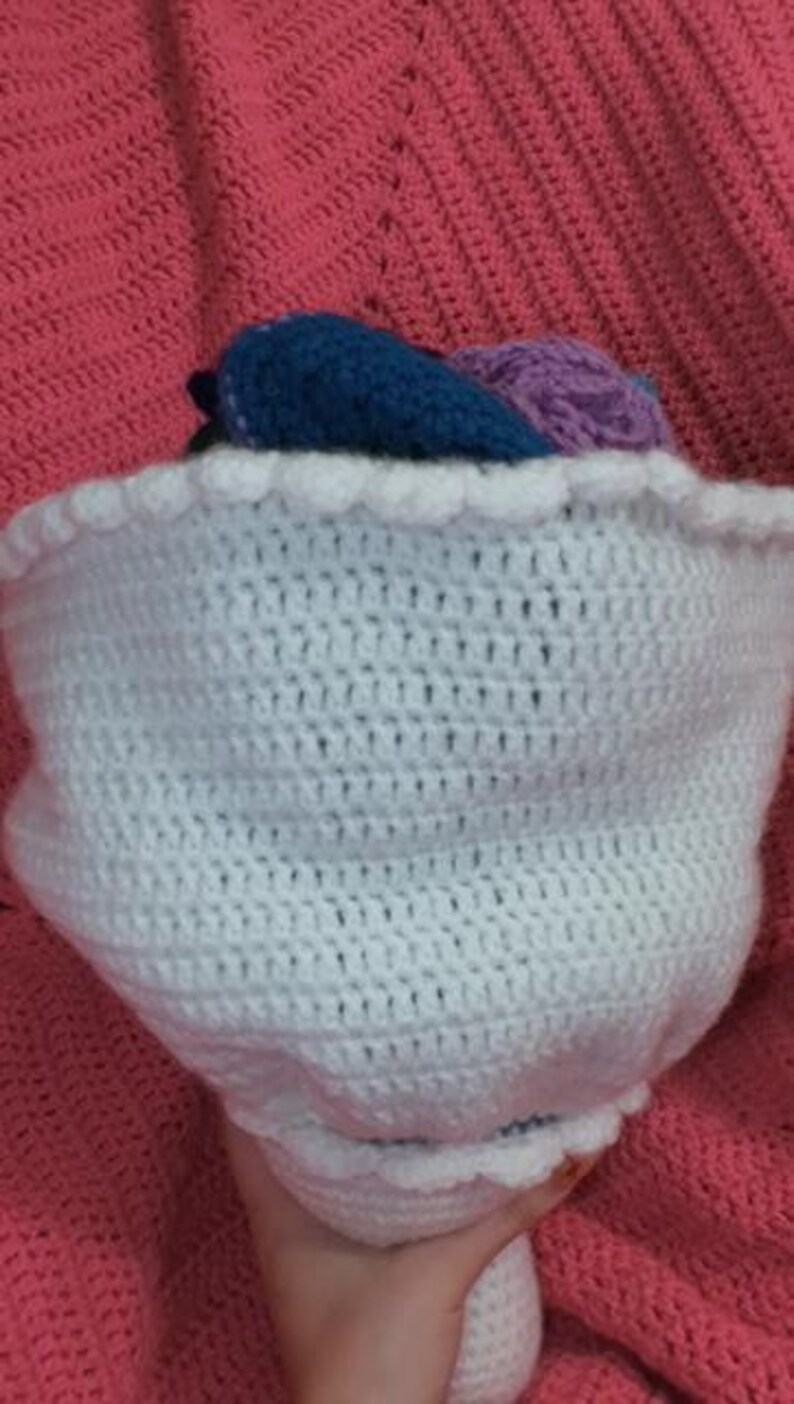 May include: A bouquet of crocheted flowers wrapped in a white crocheted paper-style sleeve with a scalloped edge. The flowers feature various colors including dark blue and light purple, all crafted with intricate yarn stitches and textures.