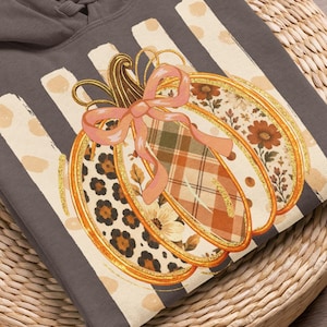 Könnte beinhalten: Ein anthrazitfarbener Kapuzenpullover mit einem grafischen Kürbis-Aufdruck und einer rosa Schleife. Das Kürbis-Design besteht aus Abschnitten mit Leopardenmuster, Karomuster und Blumenmustern vor einem Hintergrund aus vertikalen cremefarbenen Streifen.