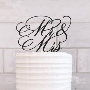 以下が含まれることがあります： 「Mr & Mrs」と黒色の筆記体で書かれたトッパーが飾られた、白い2段のケーキです。