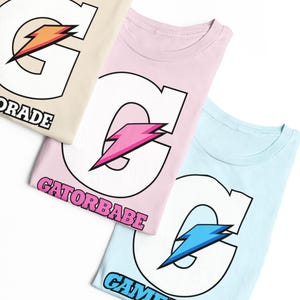 Puede incluir: Tres camisetas en beige, rosa claro y azul claro. Cada camiseta presenta una letra G blanca grande con un gráfico de rayo en naranja, rosa o azul. El texto GATORADE, GATORBABE y GAMEADE está impreso debajo de la letra G.