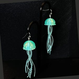 May include: A pair of teal blue jellyfish dangle earrings. The jellyfish have a translucent, slightly iridescent finish and have long, thin tentacles.