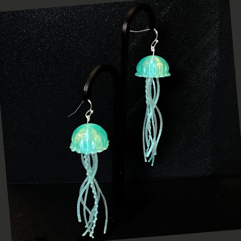 May include: A pair of teal blue jellyfish dangle earrings. The jellyfish have a translucent, slightly iridescent finish and have long, thin tentacles.