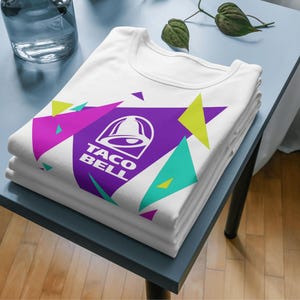Puede incluir: Una camiseta blanca con un gr&aacute;fico de tri&aacute;ngulo grande de color morado con el logotipo y el texto de Taco Bell en blanco, rodeado de formas geom&eacute;tricas m&aacute;s peque&ntilde;as en colores verde lima, magenta y turquesa, doblada sobre una superficie.