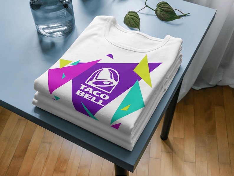 Puede incluir: Una camiseta blanca con un gr&aacute;fico de tri&aacute;ngulo grande de color morado con el logotipo y el texto de Taco Bell en blanco, rodeado de formas geom&eacute;tricas m&aacute;s peque&ntilde;as en colores verde lima, magenta y turquesa, doblada sobre una superficie.