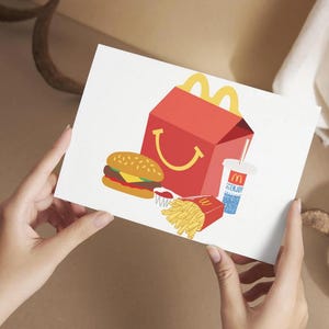 May include: A white card with a red and yellow McDonald's Happy Meal box, a hamburger, french fries, and a blue and white cup with a straw. The cup has the McDonald's logo on it.