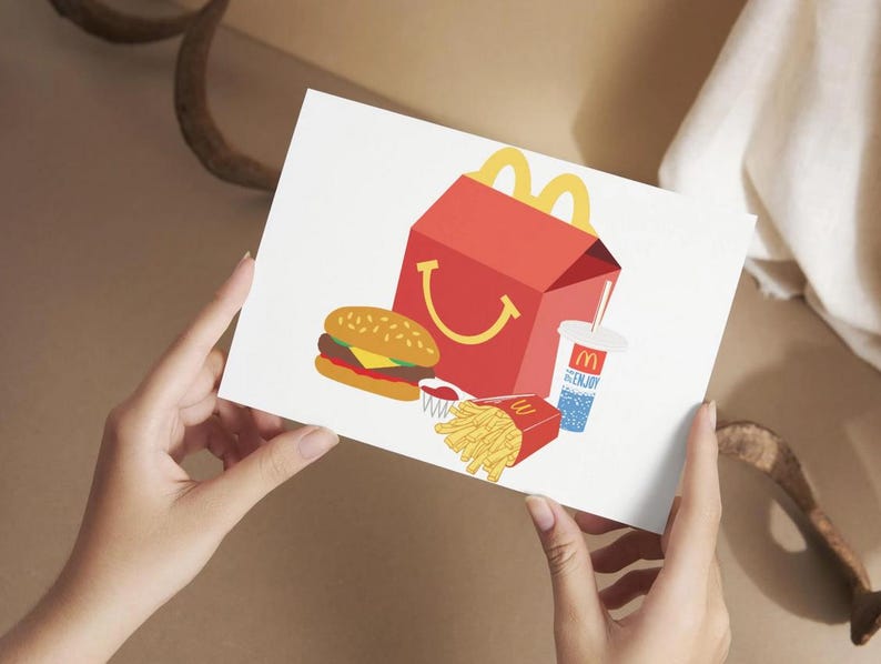 May include: A white card with a red and yellow McDonald's Happy Meal box, a hamburger, french fries, and a blue and white cup with a straw. The cup has the McDonald's logo on it.