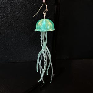 May include: A pair of green and white jellyfish earrings. The jellyfish are made of resin and have a translucent, iridescent finish. The tentacles are made of white wire and are strung with small beads.