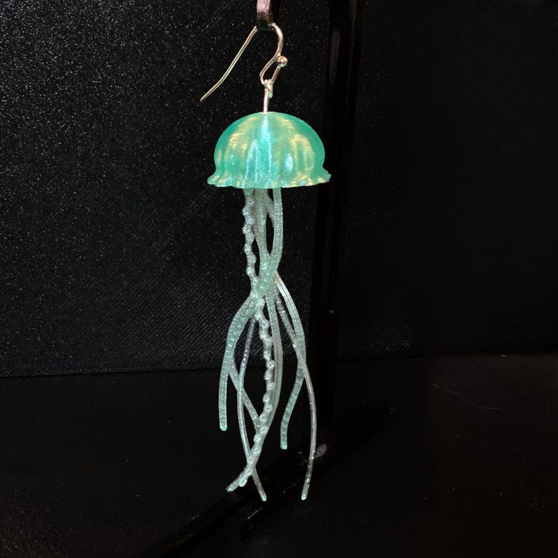 May include: A pair of green and white jellyfish earrings. The jellyfish are made of resin and have a translucent, iridescent finish. The tentacles are made of white wire and are strung with small beads.