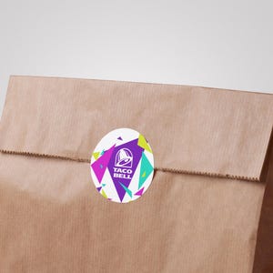 Puede incluir: Una bolsa de papel marr&oacute;n sellada con una pegatina circular que muestra el logotipo de Taco Bell en blanco sobre un fondo morado, rodeado por un patr&oacute;n geom&eacute;trico de tri&aacute;ngulos rosa brillante, verde lima y turquesa sobre una base blanca.
