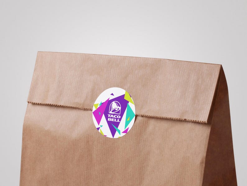 Puede incluir: Una bolsa de papel marr&oacute;n sellada con una pegatina circular que muestra el logotipo de Taco Bell en blanco sobre un fondo morado, rodeado por un patr&oacute;n geom&eacute;trico de tri&aacute;ngulos rosa brillante, verde lima y turquesa sobre una base blanca.