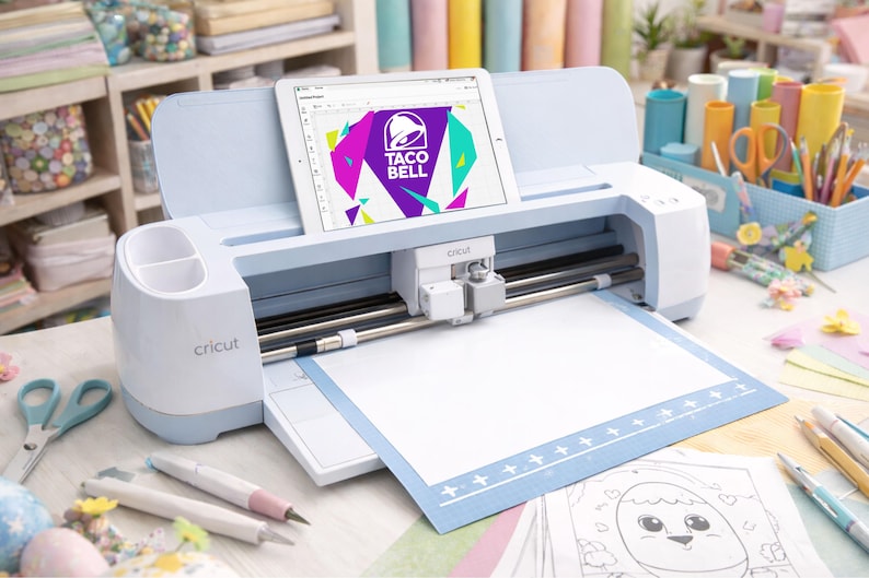 Puede incluir: Una m&aacute;quina de corte electr&oacute;nica Cricut de color azul claro con una tableta acoplada en la ranura superior que muestra un logotipo de Taco Bell. La m&aacute;quina cuenta con un carro de corte blanco, rieles met&aacute;licos y una alfombrilla azul.