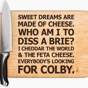 May include: A wooden cutting board with a funny cheese-themed saying: "Sweet dreams are made of cheese. Who am I to diss a brie? I cheddar the world & the feta cheese. Everybody's looking for Colby."