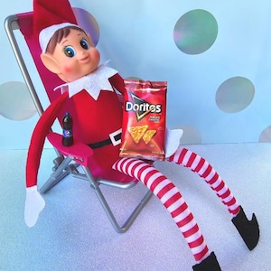 Puede incluir: Una muñeca de elfo roja y blanca sentada en una silla plegable rosa y plateada. El elfo sostiene una bolsa de Doritos con sabor a queso Nacho y una lata de refresco azul. El elfo lleva calcetines a rayas rojas y blancas.
