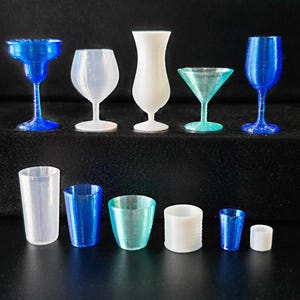 Op de afbeelding: Een verzameling van 10 heldere en blauwe glazen bekers en shotglazen. De glazen zijn in twee rijen op een zwart oppervlak gerangschikt.