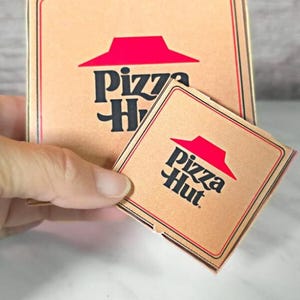 Könnte beinhalten: Zwei braune Pizzakartons mit dem Pizza Hut Logo. Die größere Schachtel hat ein rotes Dachlogo und die kleinere Schachtel hat ein rotes Dachlogo mit dem Text "Pizza Hut".